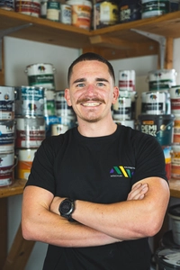 A_1_Malerbetrieb Wöschler – © Malerbetrieb Wöschler/Aurel König / Privat/Non-kommerziell Ein Porträit von Malermeister Wöschler in seiner Werkstatt. Er schaut freundlich in die Kamera. Hinter ihm stehen Farbdosen in einem Regal