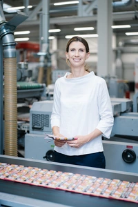 © ROSE DRUCK GMBH/Marina Terechov / Privat/Non-kommerziell Linn Rose, eine junge schlanke Frau, steht in ihrer Betriebsstätte hinter einem Fliesband und schaut freundlich in die Kamera