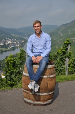 © Weingut Blesius / Privat/Non-kommerziell Florian Blesius sitzt auf einer Weintonne und lächelt freundlich in die Kamera. Im Hintergrund das Flusstal vom Rhein